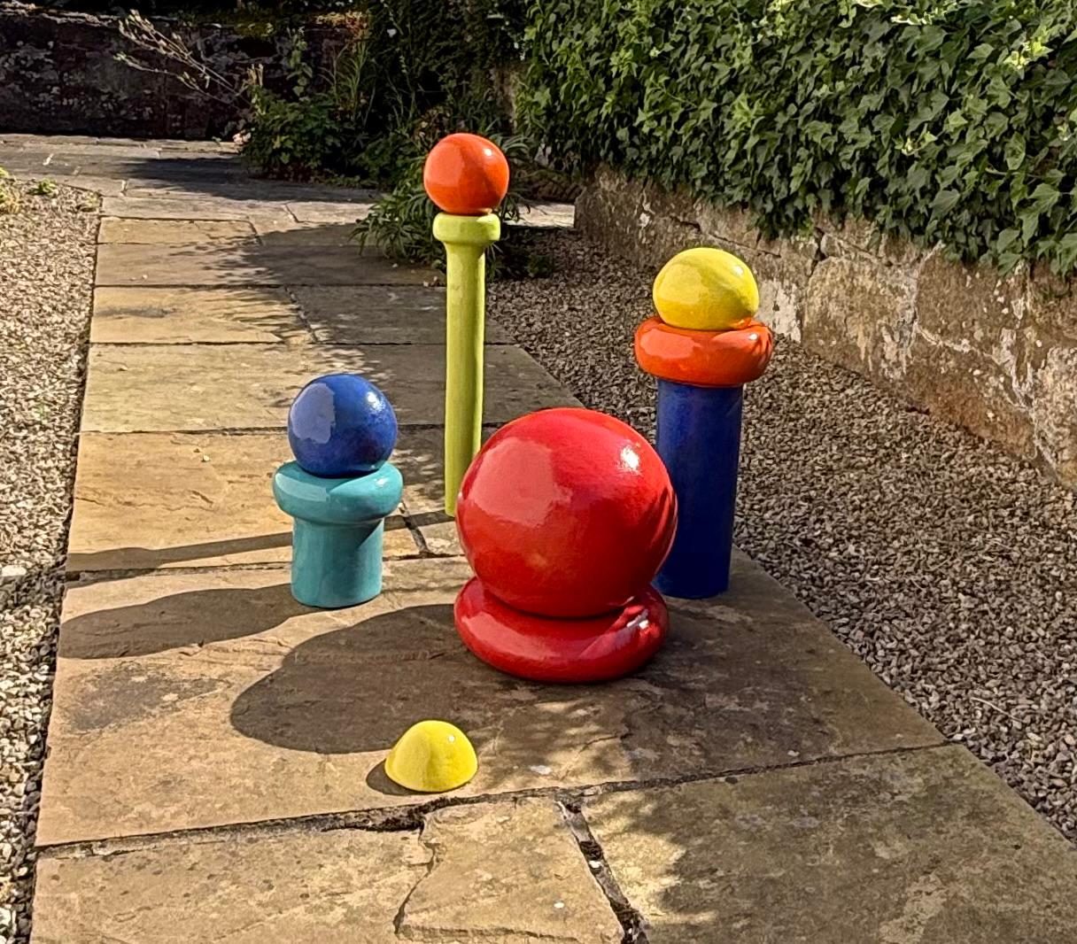 Group of brightly glazed ceramic forms — red sphere, blue and green stems, stacked orange and yellow shapes — on a garden table